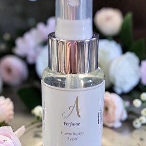 Clear glass perfume spray bottle with a translucent cap and silver collar, set among soft pink and white flowers; label shows an elegant 'A' and 'Perfume' text.
