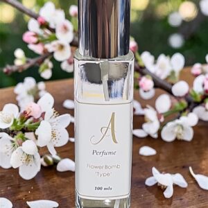 Glass perfume bottle with a metallic cap on a wooden table, surrounded by white blossoms and petals; label reads 'A Perfume Flower Bomb Type' 100 ml